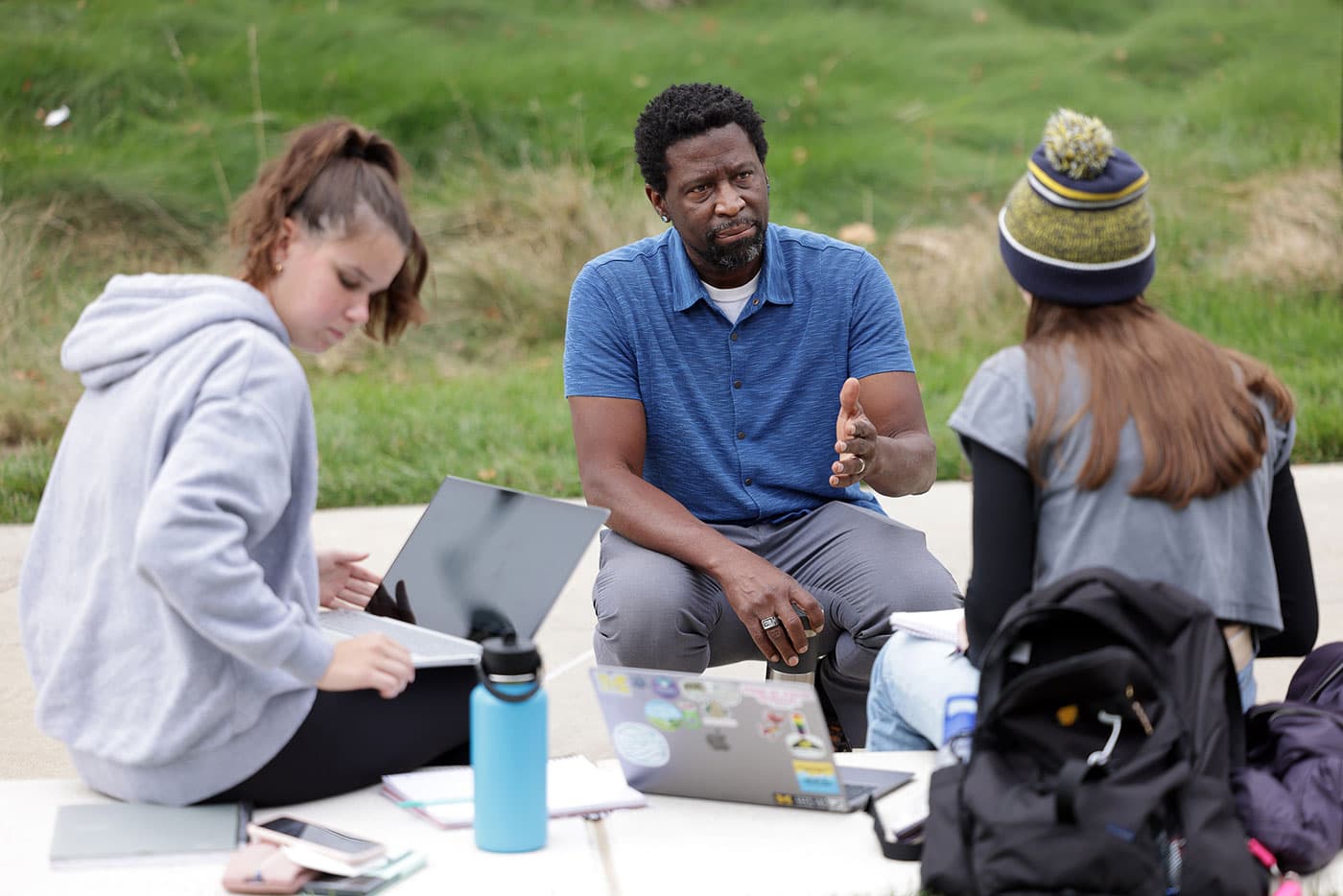 Instructor gesturing as he talks with two students seated outdoors on a sidewalk.
