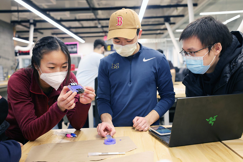 Three masked students work at a desk, designing a robot together