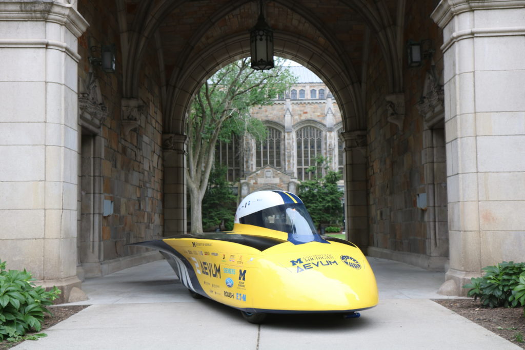 The solar car sitting under an archway on Ann Arbor campus. The car is narrow and sleek. It has a yellow finish, with blue and white splashes of color