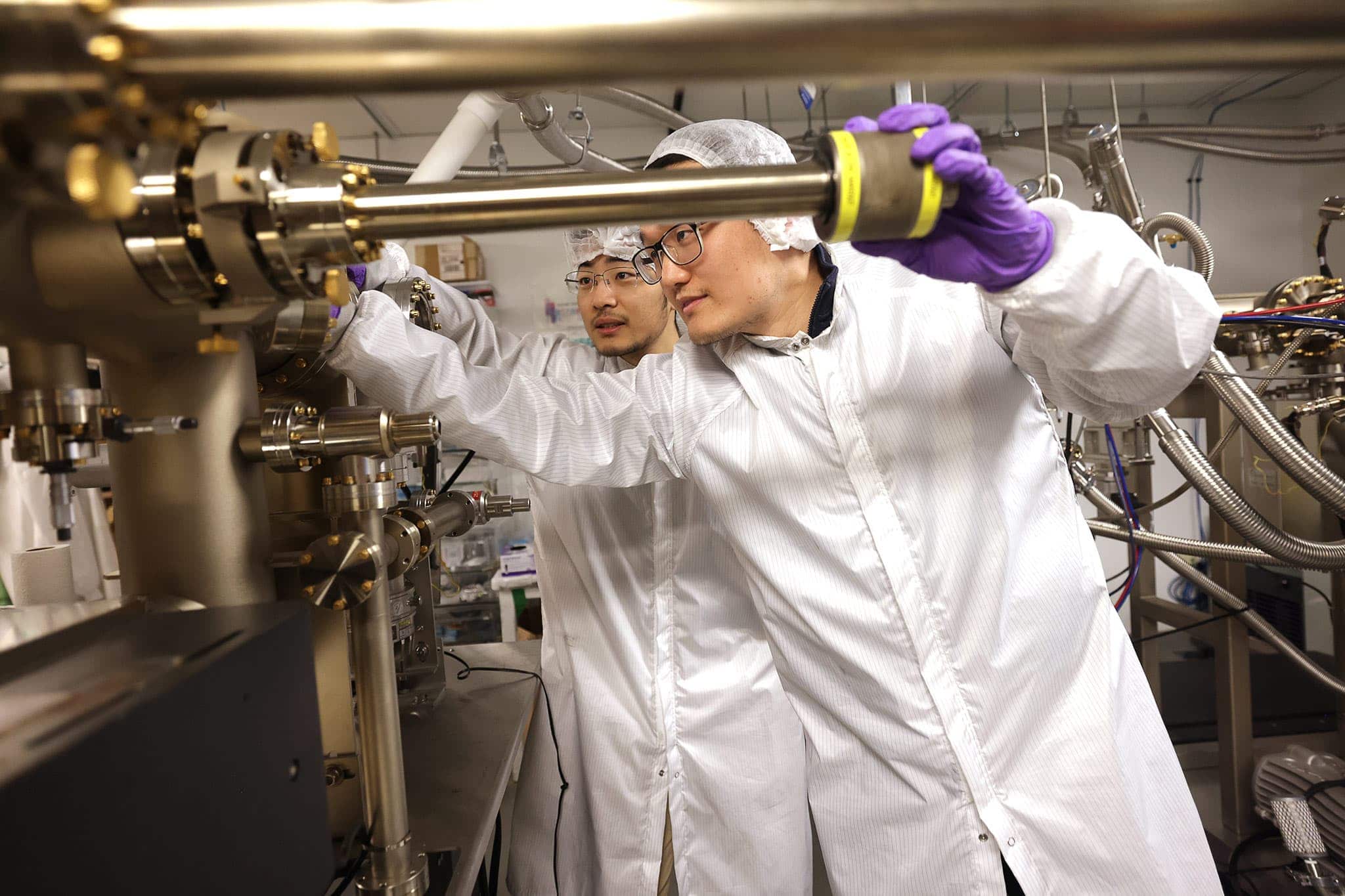 Two men in white clean room lab gear examine a large, shiny piece of machinery