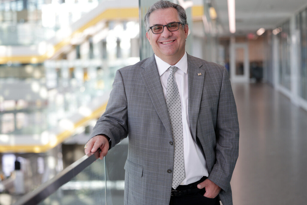 Steven Ceccio stands in a  grey suit. He rests his hand on a handrail on the second floor of the Ford Robotics Building. Below him, you can see the building atrium and the yellow stairs.