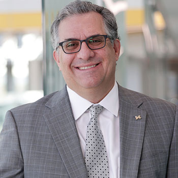 Steven Ceccio stands in a grey suit. He rests his hand on a handrail on the second floor of the Ford Robotics Building. Below him, you can see the building atrium and the yellow stairs.