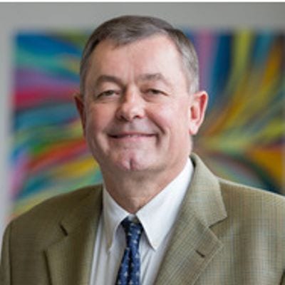 Man in tan suit with colorful pattern in background.