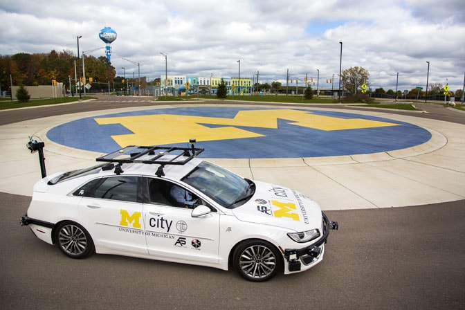 An Mcity sedan parked in a roundabout. The vehicle is white and has sensors on it. The center of the roundabout has the "block M" University logo