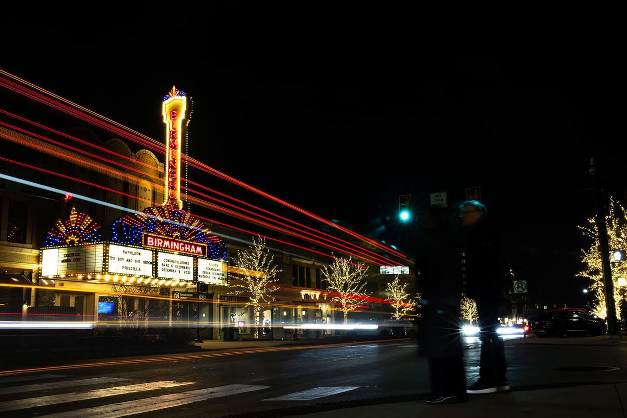 Night scene of illuminated Birmingham theater marquee with colorful lights, light trails, and blurred pedestrians.