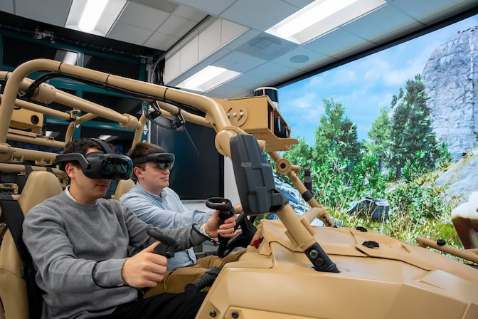 Two individuals sit in the open-framed military vehicle wearing headsets over their eyes, the virtual reality set providing total coverage while the augmented reality set resembles sunglasses hanging from a bulky visor. A large panoramic screen in front of them shows a realistic outdoor environment featuring rocky landscape with tufts of grass and conifers, creating an immersive virtual driving experience.