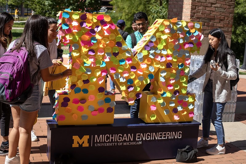 Students add colorful sticky notes to a large block M