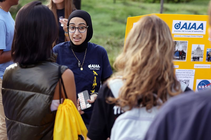 A student next to a trifold poster talks to two other students at an outdoor fair