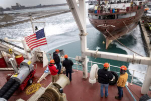 Five engineers in hard hats look out at the Nicholson Dock from the bow of a large, commercial ship, with a nearby American flag waving in the breeze overhead. The stern of another commercial ship, labeled the Herbert C. Jackson, is visible ahead of the engineers.