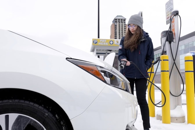 A woman, wearing a jacket and winter hat, prepares to connect an electric vehicle to a charger.