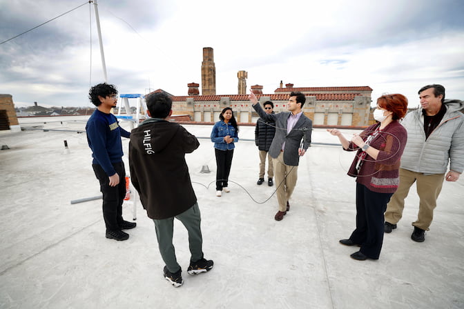 Seven people stand on a flat roof with the roofs of residential homes and a taller building with a terracotta roof in the background. A man in a blazer in the center points up at a PVC pipe with wires and ropes hanging from it while others look on. One woman holds a rope guyline coming from the PVC pipe.