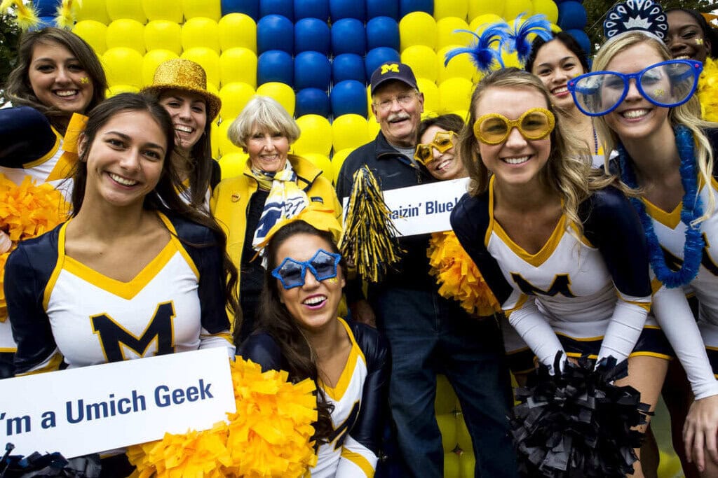 A group of cheerleaders and two adults in front of maize and blue balloons, all smiling and holding pom-poms.