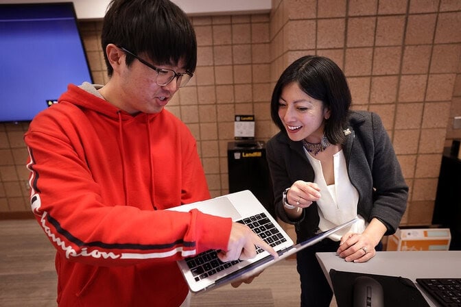 Two people discussing and pointing at a laptop in an indoor setting with a tiled wall and a blue screen in the background.