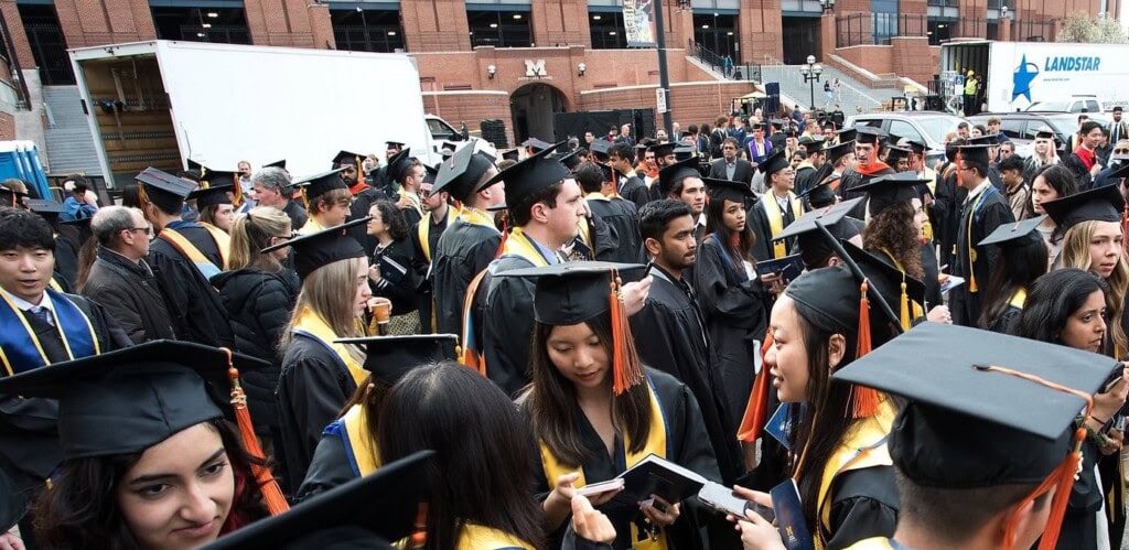 A crowd of University of Michigan College of Engineering graduates in black caps, black gowns, and orange tassels, stand shoulder to shoulder outside the Big House Stadium after a graduation ceremony.