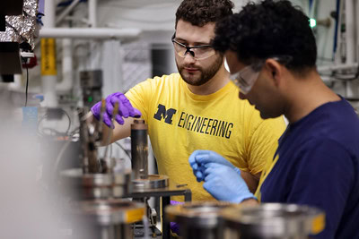  Two people working with equipment in a laboratory, wearing safety goggles and gloves. The person on the left is wearing a yellow t-shirt with "MICHIGAN ENGINEERING" written in bold letters.