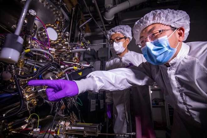 Two people in sterile protective gear work in a lab, standing before a large machine with wires, dials, and metal components. The person on the right points to a piece of equipment.