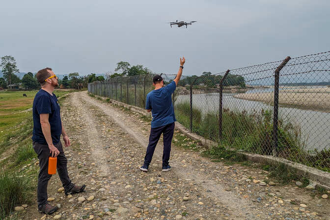 Two people on a gravel road beside a river, flying a drone.