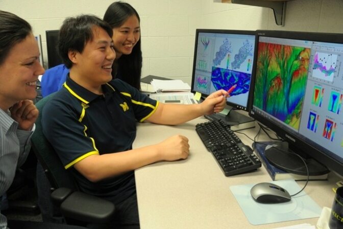 Three people discussing data visualizations on dual computer monitors.
