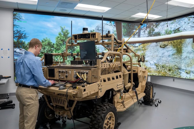 A man operates a tan unmanned vehicle with technology in a lab, against a virtual rocky landscape backdrop.