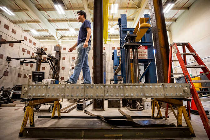 Man walking on a metal beam in an industrial lab with machinery and a red ladder.