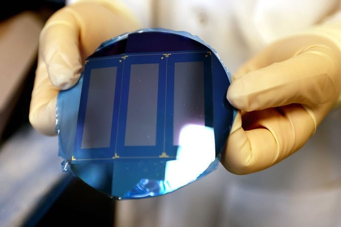 Gloved hands holding a blue, medical device chip featuring three gray rectangular patches on it and a reflective surface.