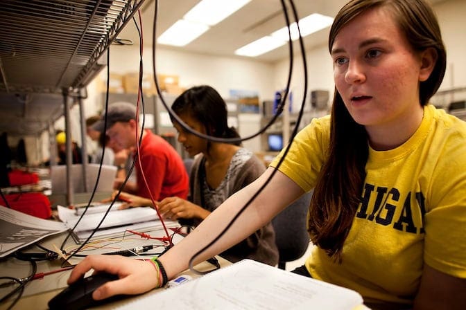 Students working with computers and electronics in a classroom, laboratory setting.The room is bright, with fluorescent lighting and shelves containing various equipment.