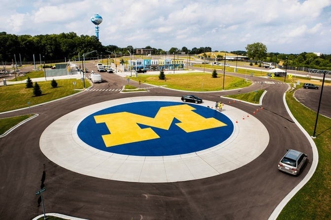 A roundabout with a large blue circle and yellow, University of Michigan block "M" in the center, surrounded by roads and greenery.