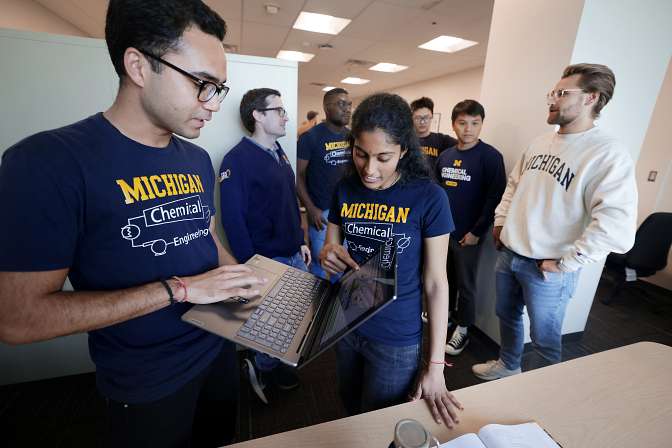Two students stand as they work together on a laptop, while other people stand gathered behind them.