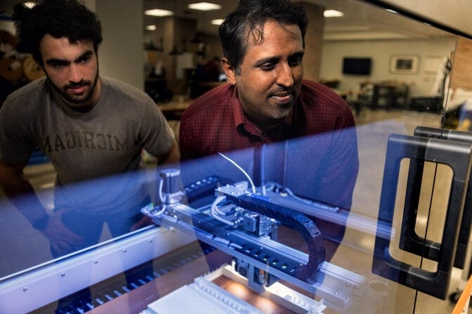 Two men are reflected in the glass of the 3D printer, which is producing a plastic structure.