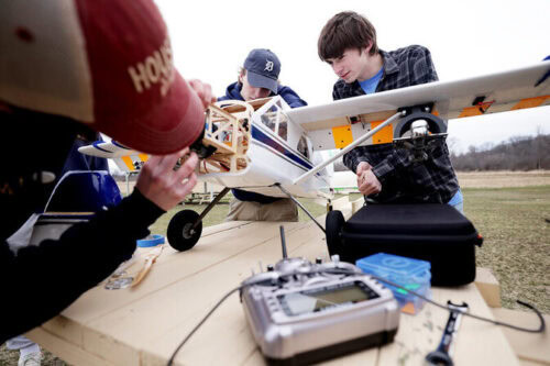Three people work on a remote-controlled airplane on a wooden table outdoors.