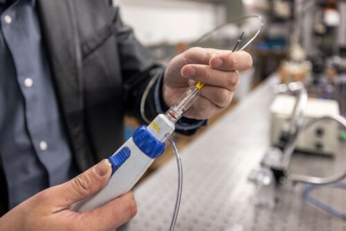 A device with a plastic handle and a metal tip is held in a researcher’s hand in a lab space.