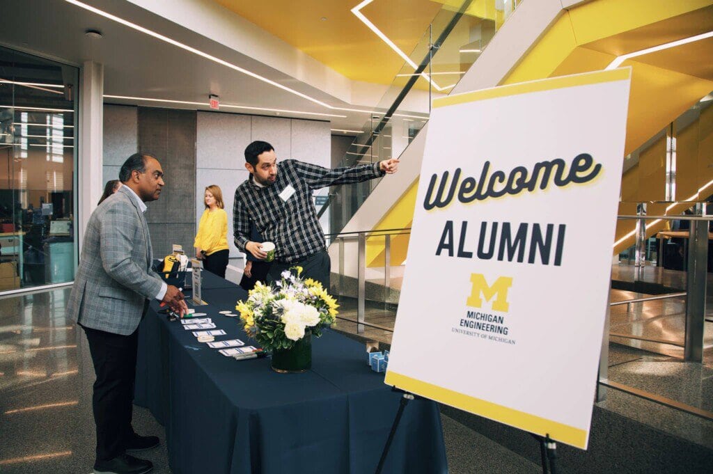 A man points to give directions to an attendant at an event. There is a sign in the foreground that reads Welcome Alumni.