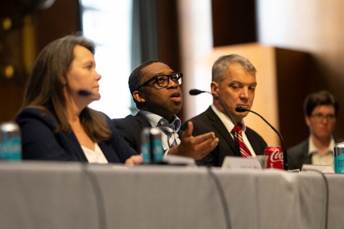 Chinedum Okwudire sits at a panel table and speaks into a microphone. 