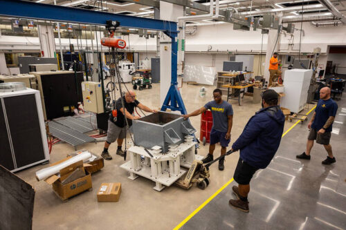 People in a workshop handling large equipment with a ceiling crane.