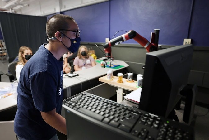 Person operating a robotic arm using a computer in front of a seated students.