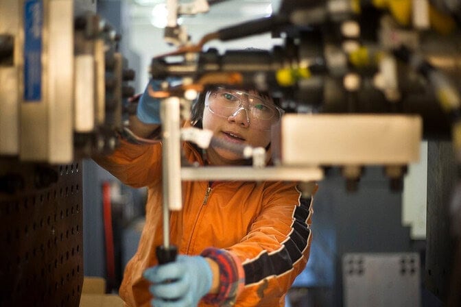 Person in a bright orange jacket and goggles adjusting machinery in a lab.