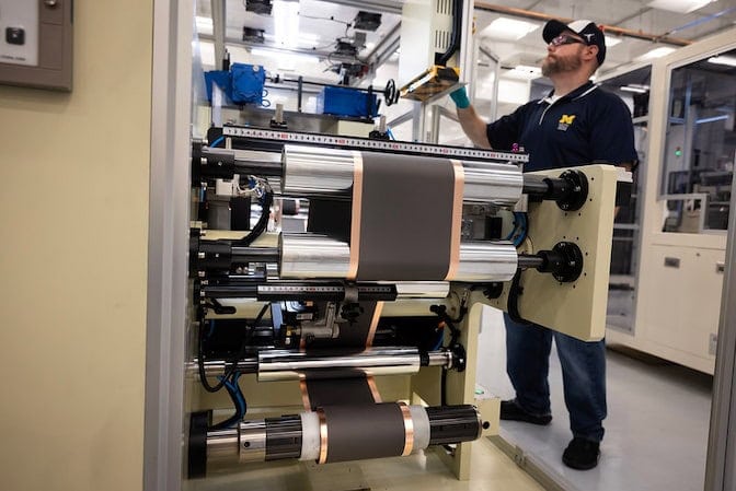 A man operates a machine featuring a roller component that is processing a dark, copper-edged material in an industrial setting.