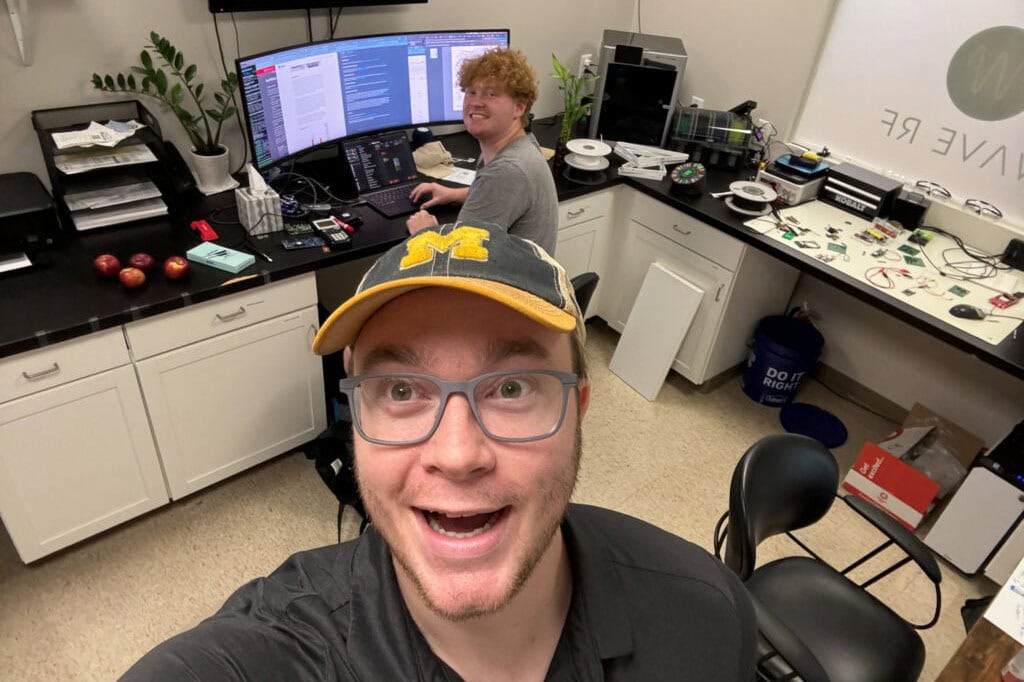 Selfie with sSmiling man in the foreground and another man sitting at a computer behind him