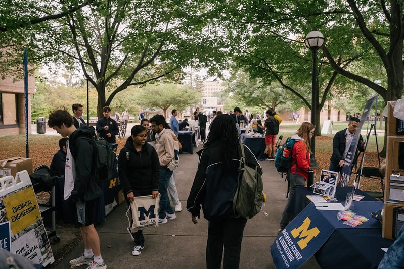 Outdoor campus walkway that is lined with trees during an openhouse event. Tables are spaced along the walk with various signs and stations for students to interact with.