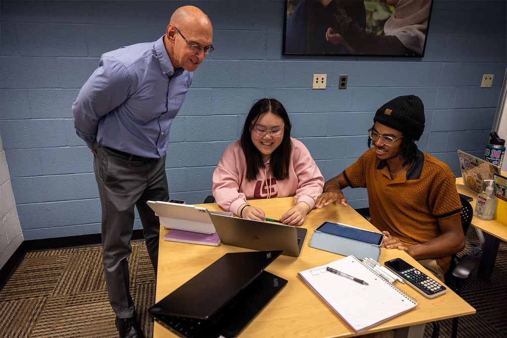 Two students and instructor gather around laptop in learning environment.