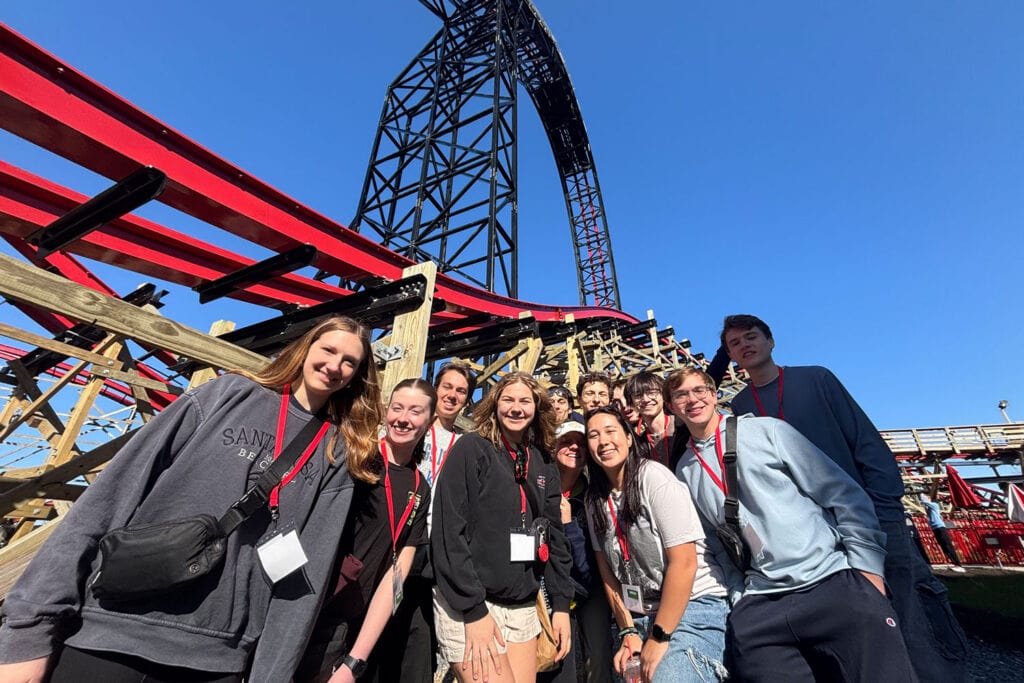 A group of 11 students pause for a group photo at a theme park. Large roller coaster track and supports are framed behind them.