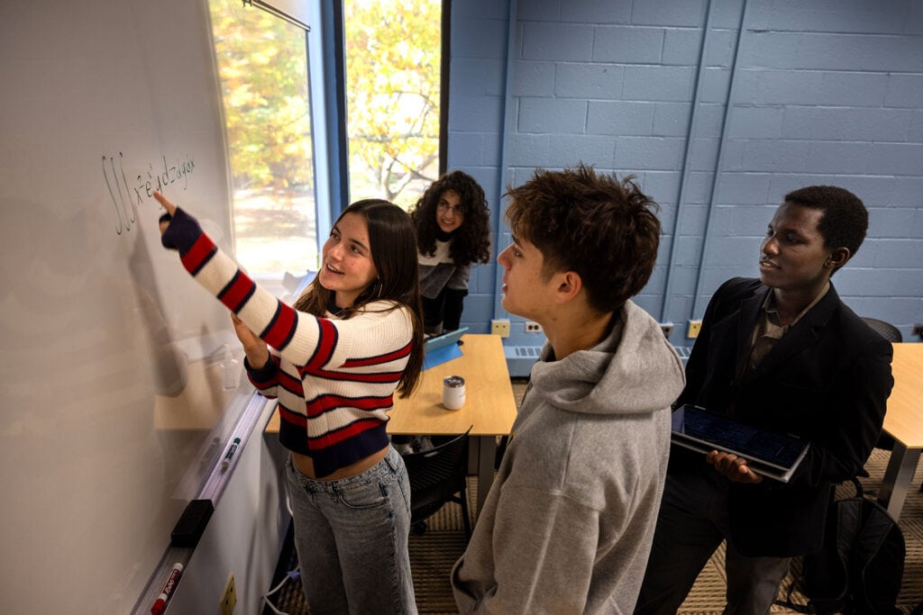 Three student gather around whiteboard and while a female points to a sentence on the board.