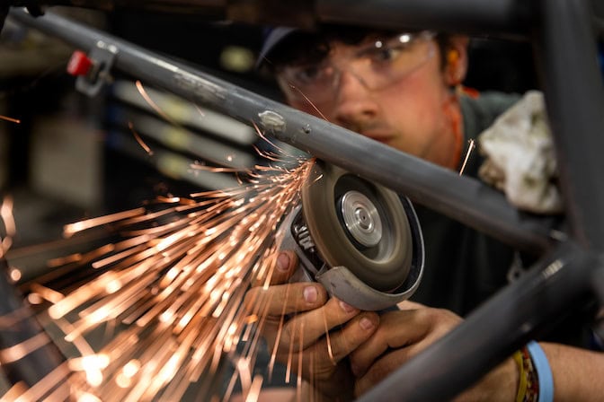 Person in safety goggles uses an angle grinder to cut a metal pipe, sending bright sparks toward the lower left.