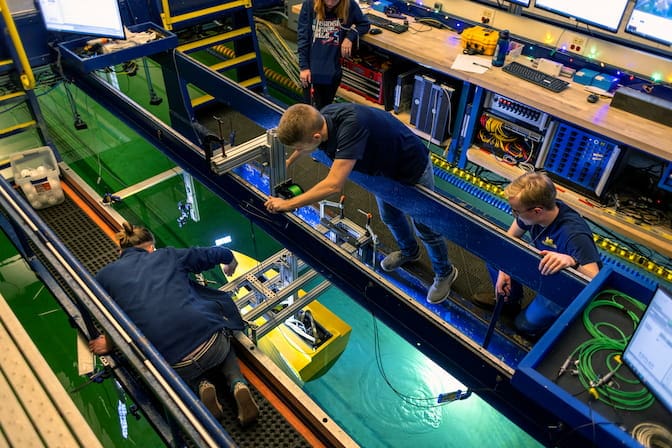 Four people stand on a platform suspended over water. Two men lean over a model ship, which is lowered into the water with a metal pole attached to a railing that runs across the center of the platform.